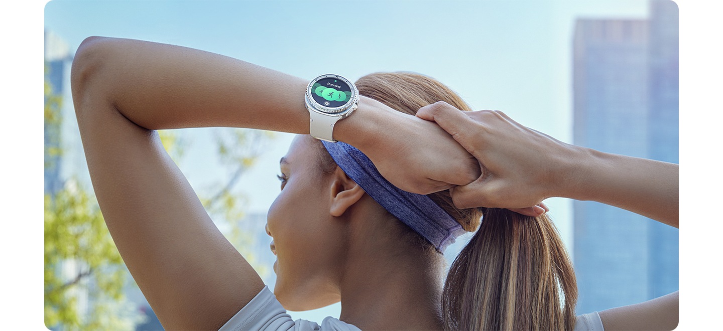 A woman wearing Galaxy Watch8 Classic paired with a White Galaxy Watch8 Sport Band stretches her arms behind her head before starting her workout in the city.