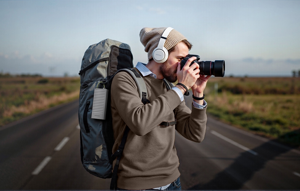 Un bărbat cu rucsac și căști face o fotografie cu un DSLR pe marginea drumului, cu un hub atașat de rucsac
