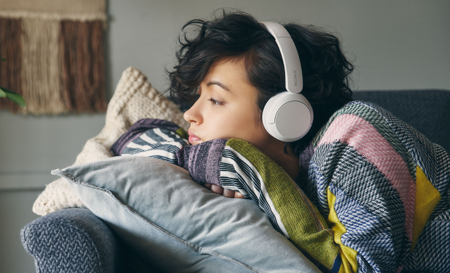 Imagine cu o femeie intinsa pe o canapea gri, cu o pereche de casti Sony WH-CH520 albe